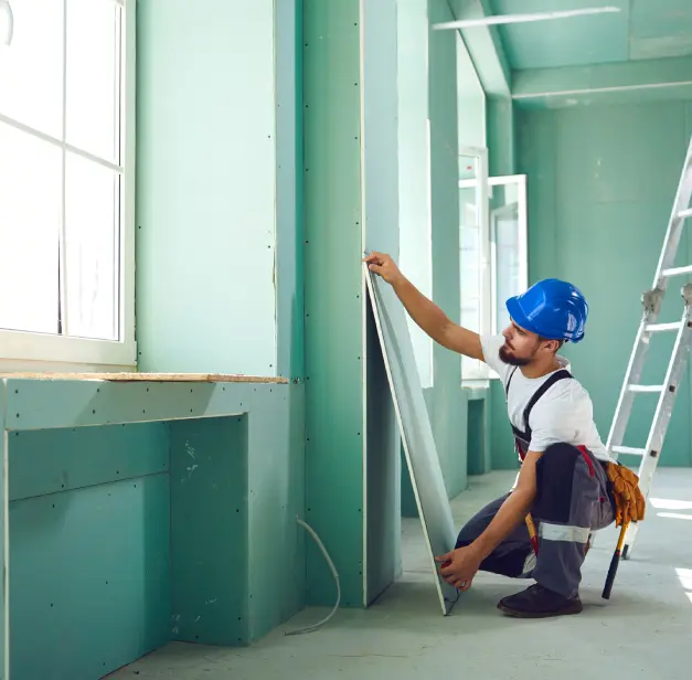 Worker in a blue hard hat installs a panel in a green room. A ladder is visible in the background.